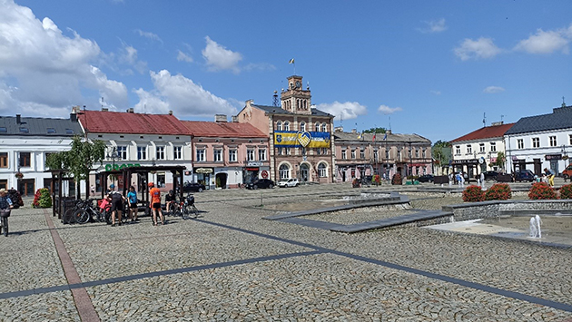miejski rynek wyłożony kostką, w tle kamienice, z lewej strony przy studni stoi grupa rowerzystów