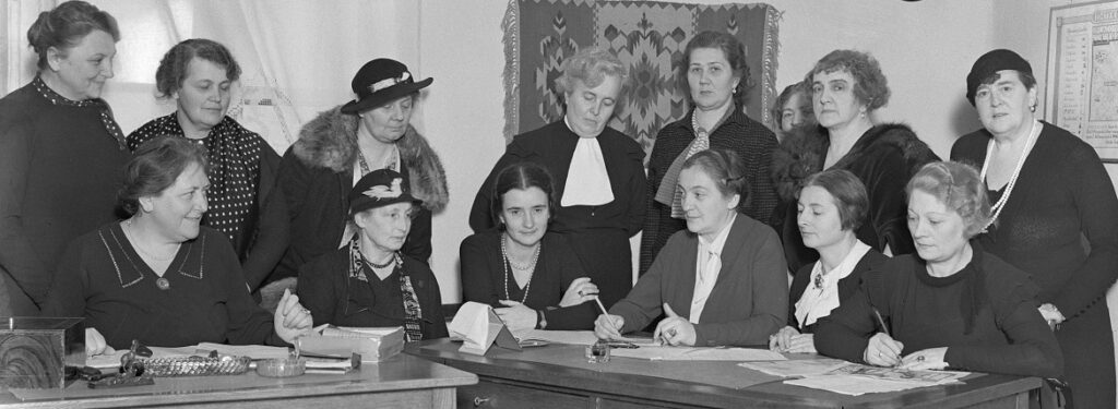 14 kobiet; część siedzi za stołem konferencyjnym; część stoi