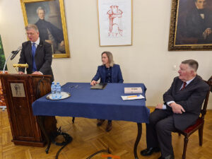 Dwie osoby siedzą przy stole, jedna stoi przy mównicy