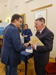 Uścisk dłoni dwóch osób, jedna z nich trzyma książkę, w tle widoczna trzecia osoba