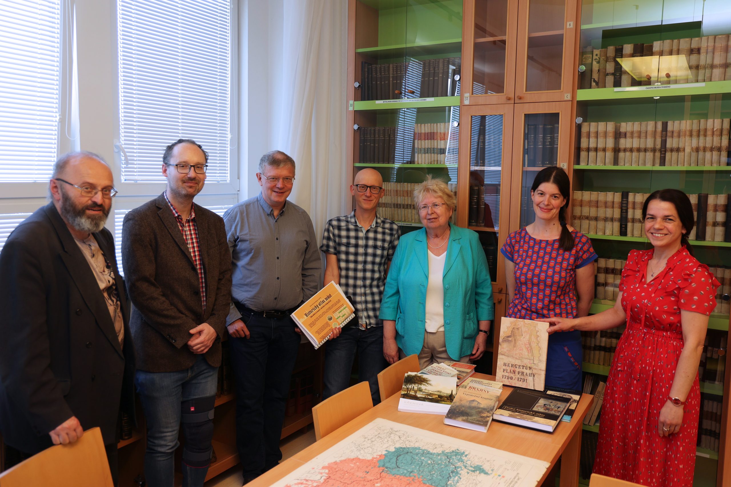 7 osób stojących przy stole; na stole leżą mapy i książki; po lewej za plecami jest okno, a z prawej oszklony regał z książkami.